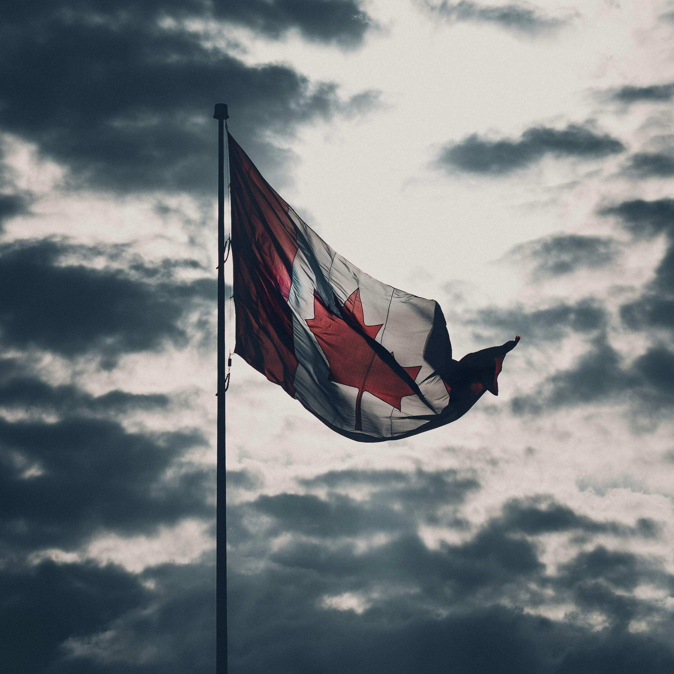 Home A dramatic view of the Canadian flag waving against a backdrop of cloudy skies, highlighting national pride.